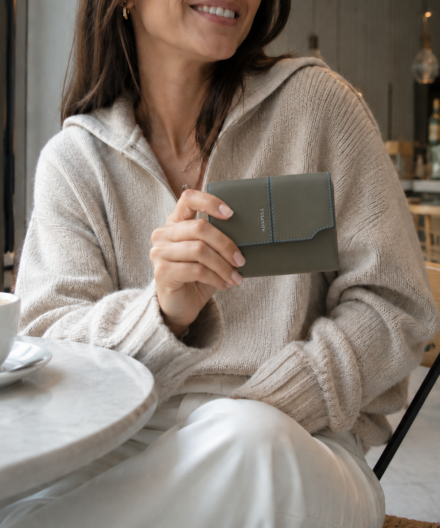 Mujer sosteniendo una cartera pequeña Atelier en color Verde Musgo en una cafetería, mostrando la vista frontal con solapa, pespuntes de contraste y logo Adapell.