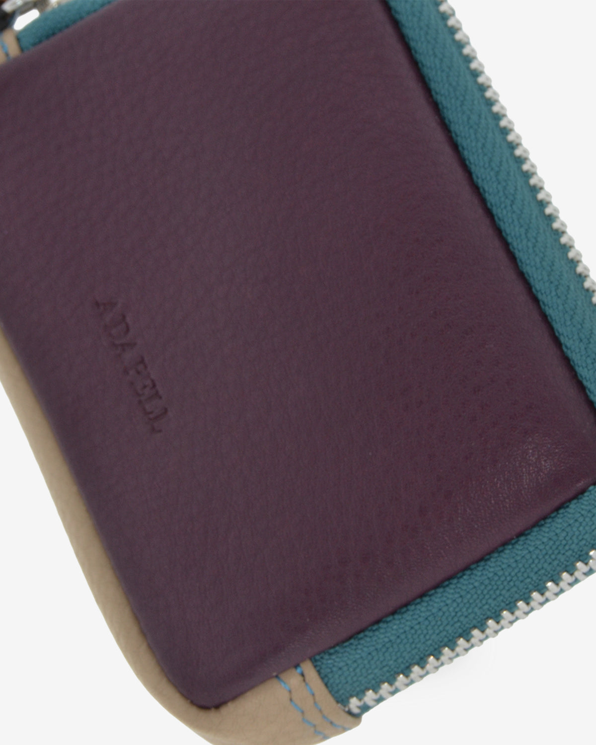 Detalle del monedero Atelier en color Berenjena con textura de la piel, logo Adapell grabado, borde azul verdoso y cierre metálico de cremallera.