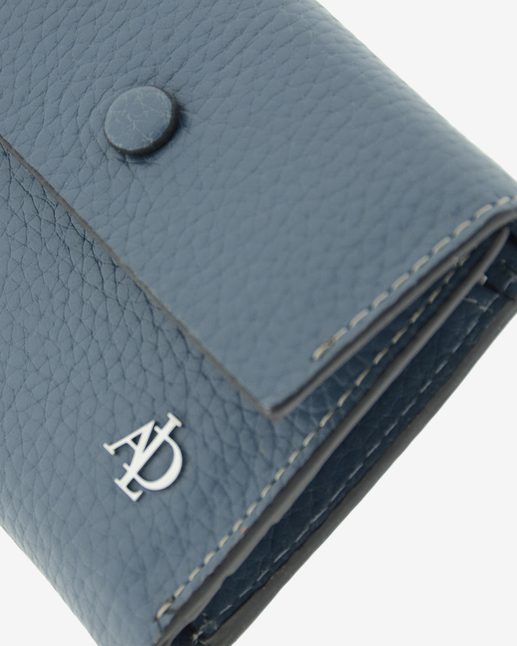 Detalle frontal de la cartera pequeña Nude en color Azul piedra con solapa, botón, costuras, textura de la piel y logo Adapell.