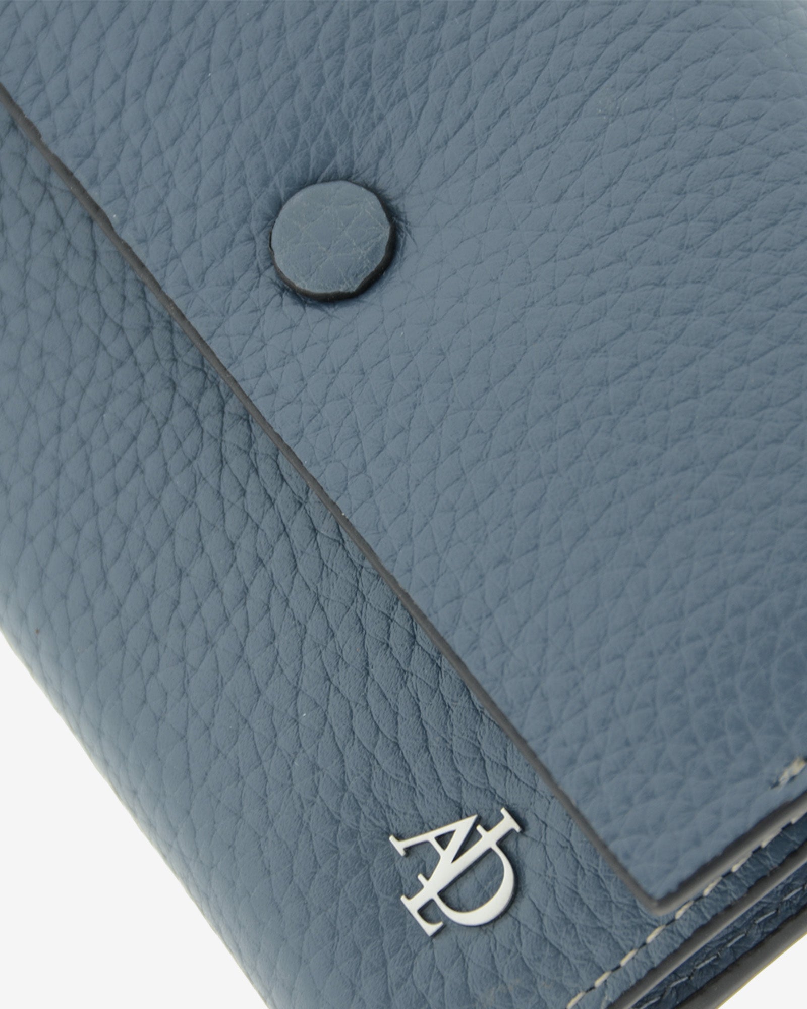 Detalle de la solapa con botón a presión, la textura de la piel y el logo metálico Adapell de la cartera Nude en color Azul Piedra.